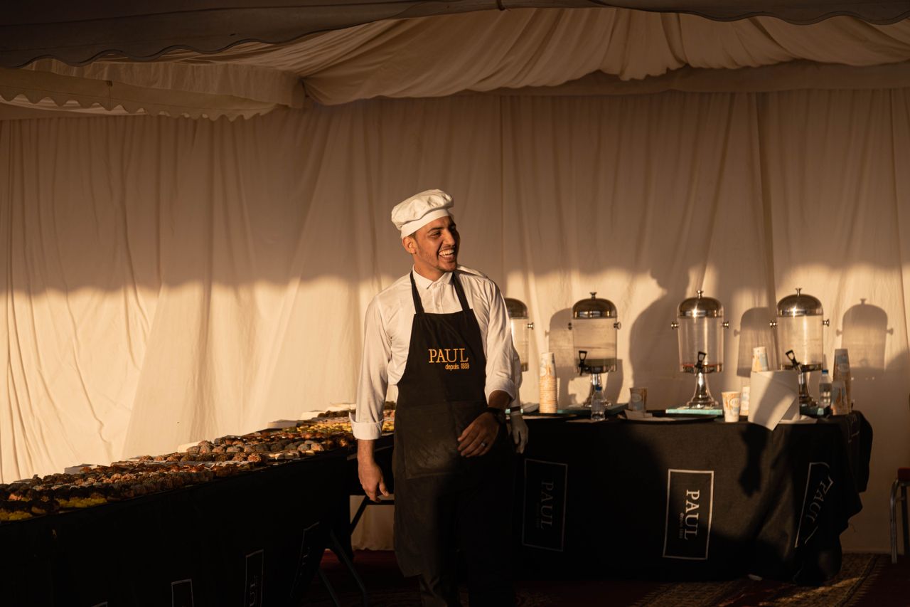 services-04 A smiling chef in an apron stands at a buffet table with pastries, creating a welcoming atmosphere.