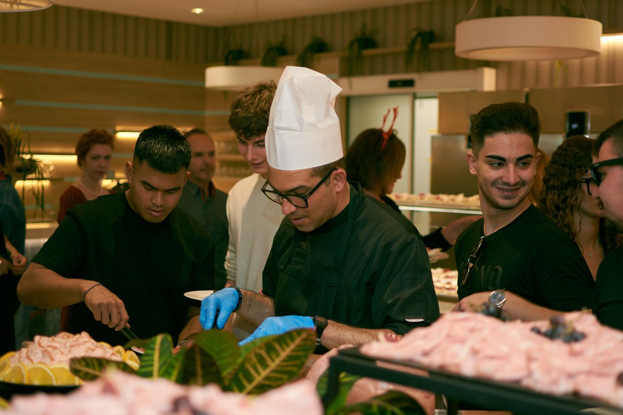 about-us A chef in glasses serves food to guests at a lively indoor buffet event.
